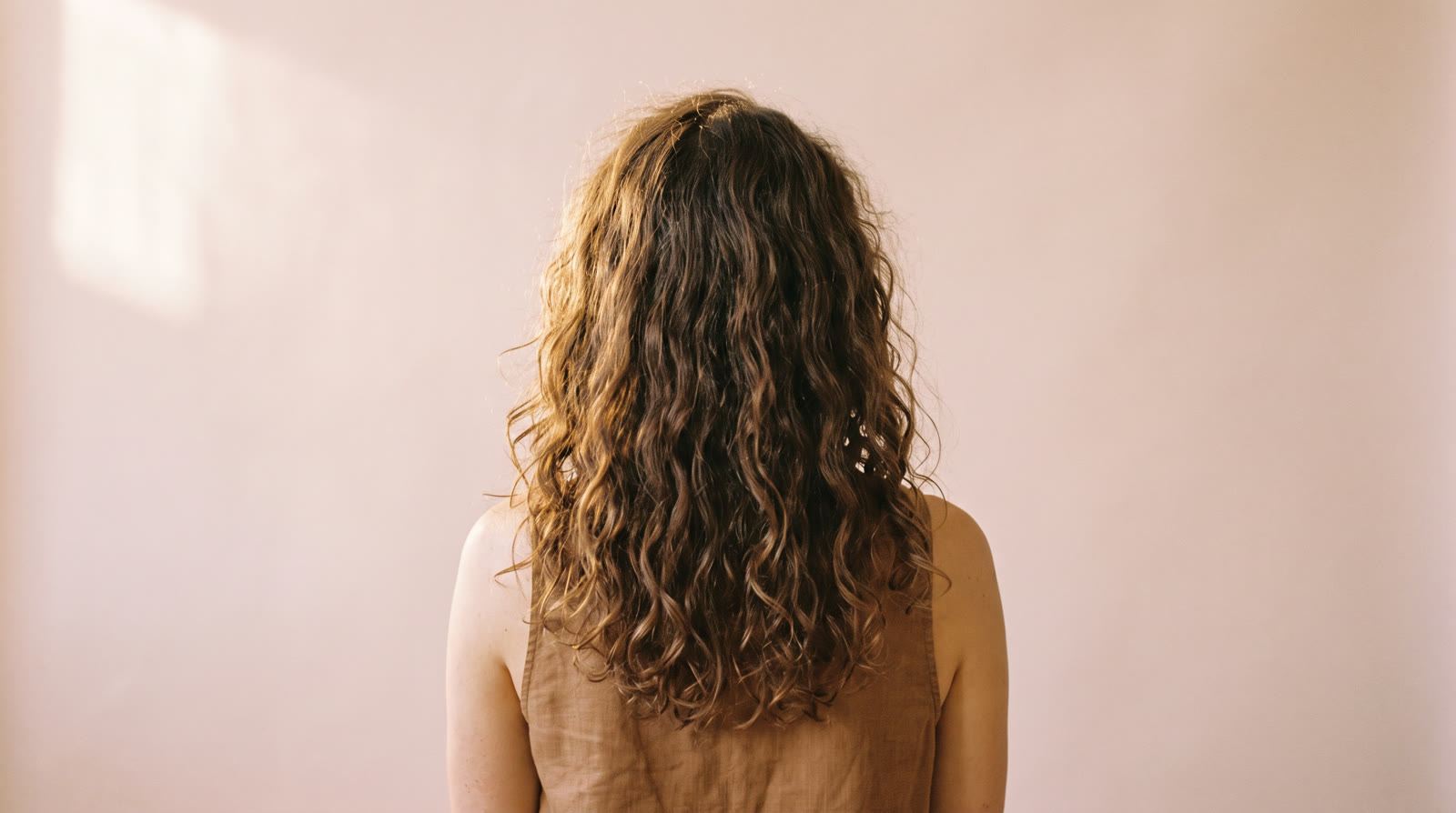 The back of a woman's head, long natural curls at rest against a pale blush studio backdrop